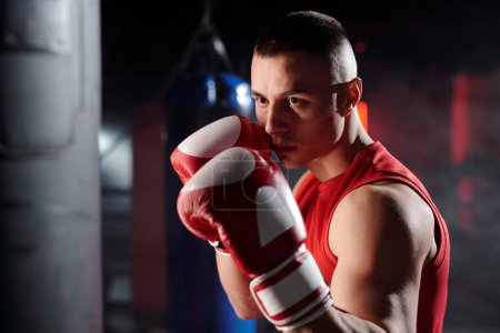 Foto de Hombre musculoso joven mirando saco de boxeo antes de golpearlo mientras está parado frente a la cámara durante el entrenamiento de boxeo en el gimnasio - Imagen libre de derechos
