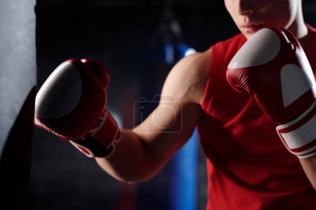 Recorte de tiro de atleta joven en ropa deportiva roja pateando saco de boxeo durante el entrenamiento deportivo mientras está de pie delante de la cámara en el gimnasio
