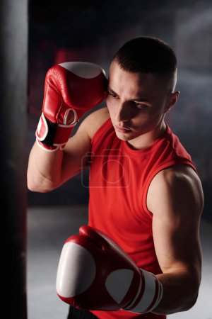 Téléchargez les photos : Jeune boxeur masculin sérieux en gilet rouge et gants de boxe regardant sac de boxe pendant l'entraînement tout en se préparant pour la compétition - en image libre de droit