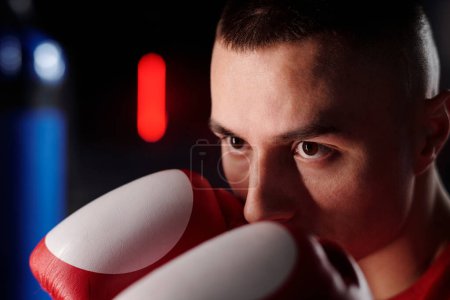 Face of young male boxer looking forwards and keeping hands in boxing gloves by nose while standing in front of punching bag