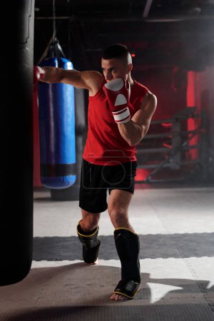 Retrato completo del joven atleta en ropa deportiva parado en el ring de boxeo y golpeando el saco de boxeo mientras entrena antes de la competición