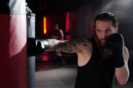 Téléchargez les photos : Homme musclé barbu avec des tatouages sur le bras et la poitrine debout devant le sac de boxe et le frapper pendant l'entraînement dans la salle de gym ou l'anneau de boxe - en image libre de droit