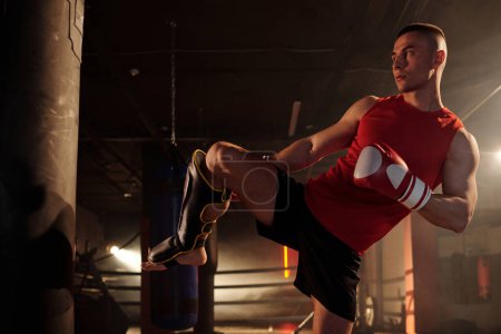Boxeador joven en ropa deportiva roja practicando patada baja mientras golpea el saco de boxeo con la pierna derecha en protectores durante el entrenamiento