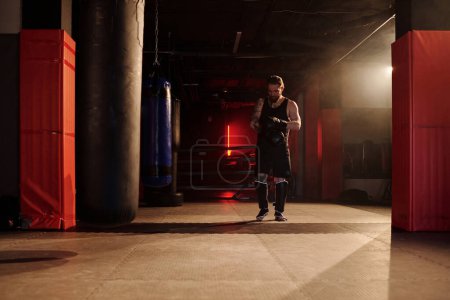 Téléchargez les photos : Plan long de jeune athlète masculin en vêtements de sport noirs marchant le long de la salle de gym avec des sacs de boxe et mettant des gants de boxe sur les mains - en image libre de droit