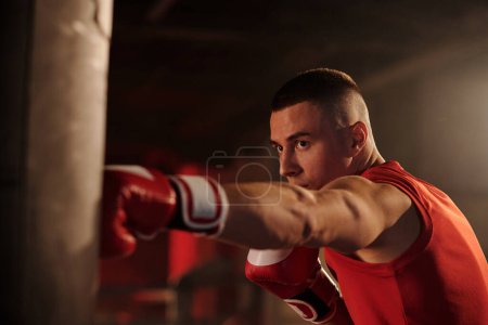 Foto de Joven hombre musculoso en guantes de boxeo rojos pateando la caja de boxeo durante el entrenamiento mientras practica ejercicios básicos antes de la primera ronda - Imagen libre de derechos