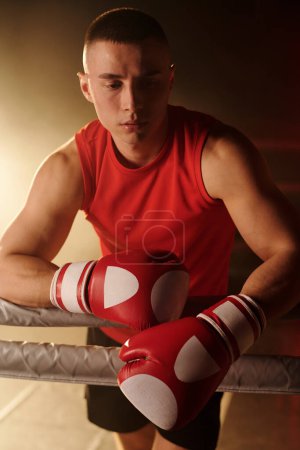 Young tired male boxer in red gloves standing on boxing ring and having break between rounds or after hard training or competition