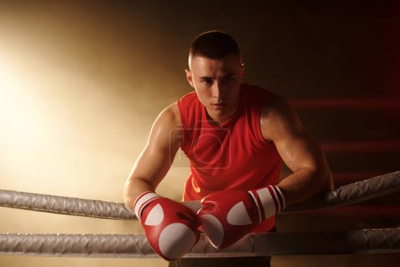 Young serene man in red vest and boxing gloves standing on ring and looking aside while having break between rounds