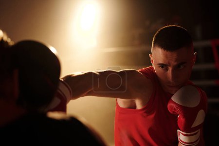 Jeune homme musclé en tenue de sport rouge et gants de boxe regardant son rival tout en le frappant à la tête lors d'une ronde sur anneau