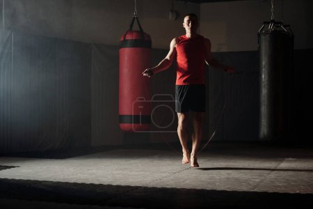 Young barefoot sportsman in red vest and black shorts jumping with skipping rope on the floor of gym with two punching bags