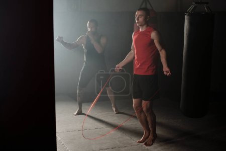 Téléchargez les photos : Lumière tombant de la fenêtre du gymnase sur deux jeunes athlètes masculins pratiquant des sauts avec corde à sauter et des techniques de boxe avec sac de boxe - en image libre de droit