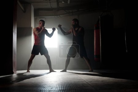 Two backlit male figures with clenched fists standing in front of one another and practicing basic sparring exercises during training