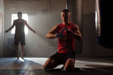 Jeune homme en gilet rouge et short noir debout sur les genoux sur le sol de la salle de gym et enveloppant la main avec bande élastique contre le gars actif