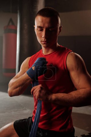 Jeune homme musclé en tenue de sport rouge assis sur le sol de la salle de gym devant la caméra et enveloppant la main avec une bande élastique avant l'entraînement