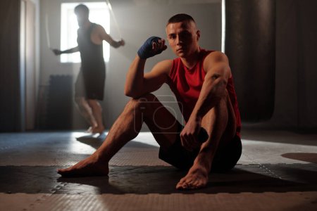 Photo for Young barefoot athlete in sportswear sitting on the floor of gym and looking at camera against guy jumping with skipping rope - Royalty Free Image