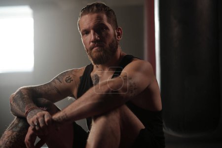 Young bearded man in sportswear looking at camera while sitting in gym and having break after hard training or boxing round
