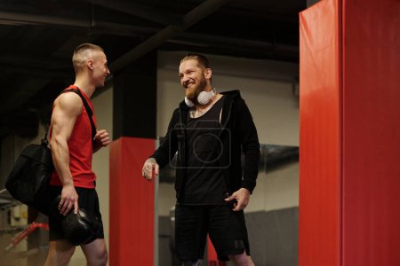 Two young cheerful male athletes in sportswear looking at one another during conversation while standing in gym before training