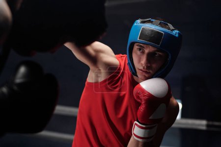 Joven boxeador con casco protector mirando a su rival y pateándolo durante el ataque o la defensa mientras practica la técnica de combate