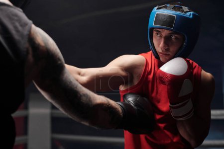 Téléchargez les photos : Main en gant de boxe noir d'athlète brutal musclé avec des tatouages donnant un coup de pied à son rival en vêtements de sport rouges et casque de protection pendant l'entraînement - en image libre de droit
