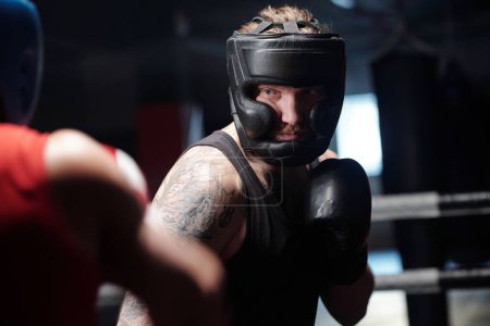 Téléchargez les photos : Jeune boxeur homme en gilet noir, écran protecteur et gants de boxe debout sur l'anneau devant son rival et le forçant - en image libre de droit