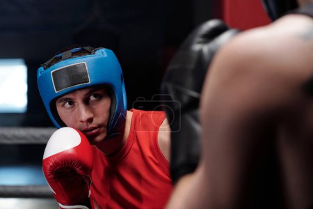 Joven atleta masculino serio en escudo azul protector y guantes de boxeo rojos de pie delante de su rival en el ring y preparándose para el ataque