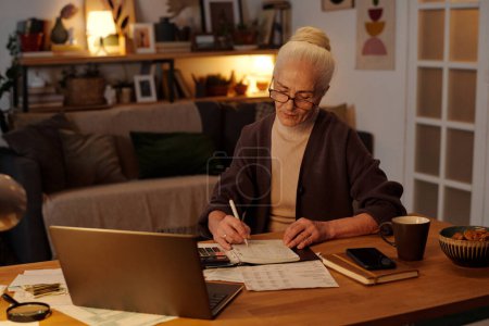 Foto de Mujer mayor en gafas graduadas y ropa casual sentada junto a una mesa de madera en la sala de estar y anotando sumas pagadas en bloc de notas o copybook - Imagen libre de derechos