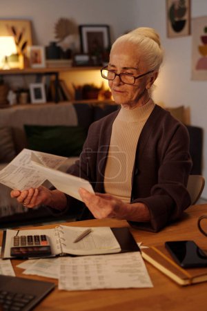 Aged serious woman with white hair sitting by table in home environment and looking through paid and unpaid financial bills