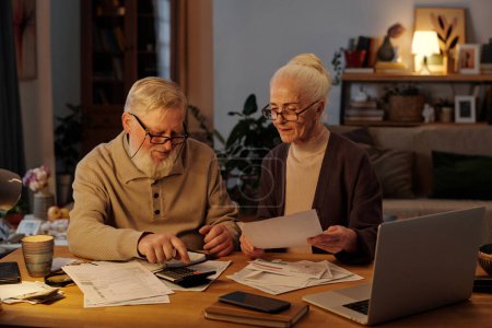 Senior man with beard pressing button of calculator while sitting next to his wife and checking sums in paid and unpaid financial bills