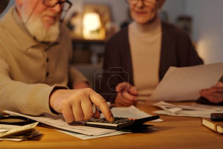 Senior man pressing calculator button while sitting by table next to his wife with financial bill in hands and both checking data in documents