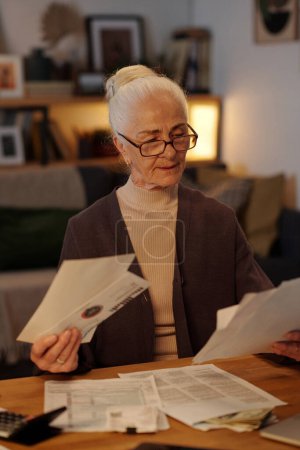 Modern elderly woman with white hair looking through financial bills and other post while sitting by table in front of camera at home