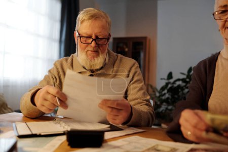 Photo for Serious mature man with white beard holding financial document with housing payment sums and looking through information - Royalty Free Image
