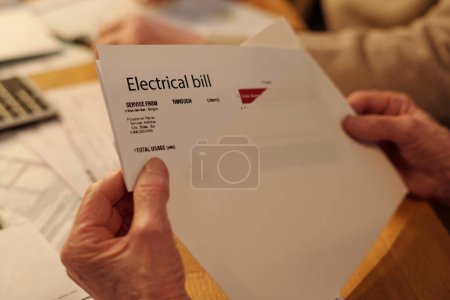 Hand of unrecognizable elderly woman taking folded electrical bill out of white envelope while sitting by table and checking payment documents