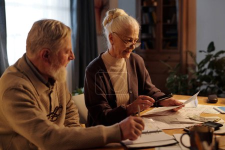 Ältere Frau in Freizeitkleidung und Brille hält Lupe über Geldschein und liest ihn, während sie neben ihrem Mann sitzt