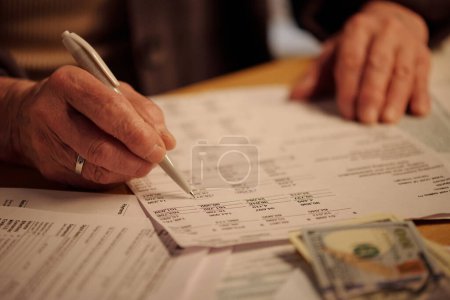 Photo for Hand of senior female owner of house or flat touching financial bill with pen while looking through and checking unpaid sums - Royalty Free Image
