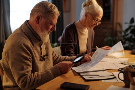 Seitenansicht eines betagten Mannes mit Lupe, der durch Finanzrechnungen blickt, während seine Frau neben ihm sitzt und Daten in Dokumenten prüft