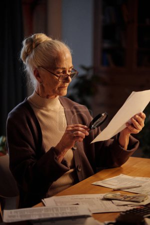 Serious aged woman in eyeglasses and casual attire sitting by table at home and reading financial document through magnifying glass