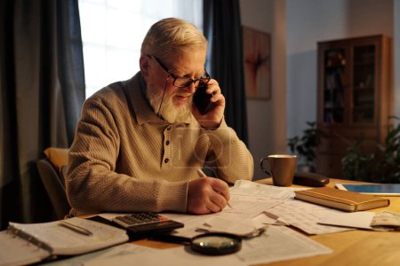 Modern senior man with white beard speaking to manager of housing service on mobile phone while specifying information
