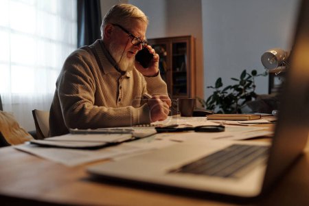 Photo for Serious senior man in eyeglasses looking through financial bill while sitting by table at home and checking information on mobile phone - Royalty Free Image