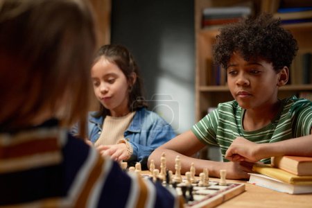 Serious African American schoolboy in striped t-shirt thinking of next move and looking at chess board with pawns and other figurines