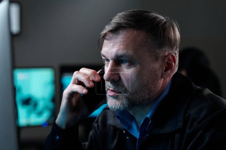 Foto de Confident mature male officer with smartphone by ear looking at computer screen while watching cctv and reporting situation - Imagen libre de derechos