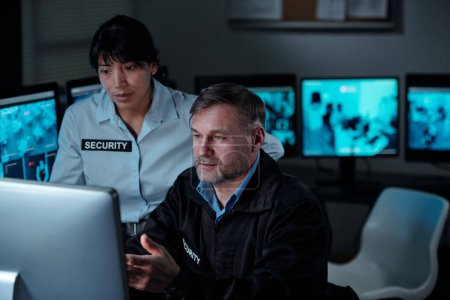 Foto de Young female and mature male security guards in uniform looking at computer screen while one of them explaining detail of video - Imagen libre de derechos