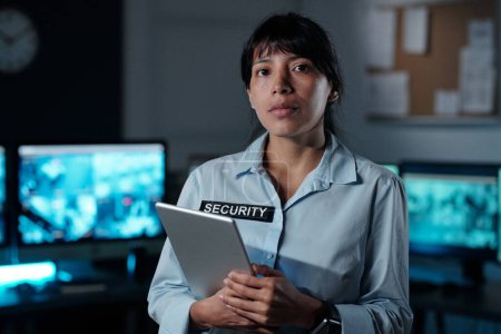 Foto de Young serious Hispanic female officer of surveillance room with tablet in hand looking at camera while standing against computer monitors - Imagen libre de derechos