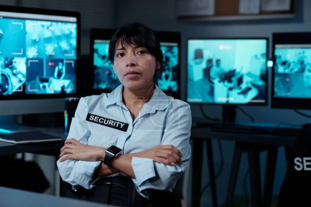 Photo for Serious young female security guard in uniform keeping arms crossed by chest and looking at camera against row of computer monitors - Royalty Free Image