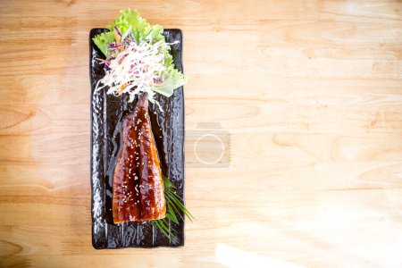 Laissez-vous tenter par les saveurs exquises de l'anguille japonaise grillée servie sur une assiette noire, magnifiquement présentée sur une table en bois.
