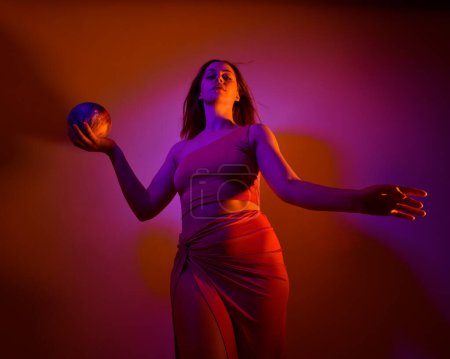 Photo for Low angle portrait of pretty girl wearing pink outfit, gestural arm poses reaching out as if casting a spell, colourful neon gel lighting, isolated on studio background. - Royalty Free Image
