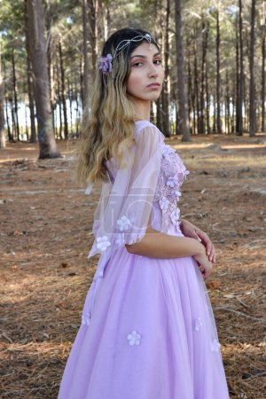 Close up portrait of beautiful young blonde model wearing a purple princess fantasy ball gown with flower crown diadem.Pine forest location background with golden lighting.