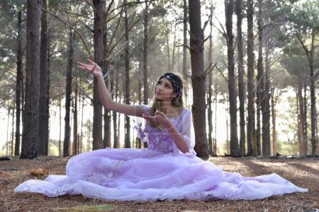 Fun length  portrait of beautiful young blonde model wearing a purple princess fantasy ball gown with flower crown diadem. Sitting pose in pine tree forest location background with golden lighting.
