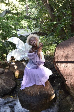 Retrato de una hermosa mujer africana con traje de fantasía púrpura, alas mágicas de hadas y corona de flores afro, vagando por la ubicación del bosque con iluminación natural.