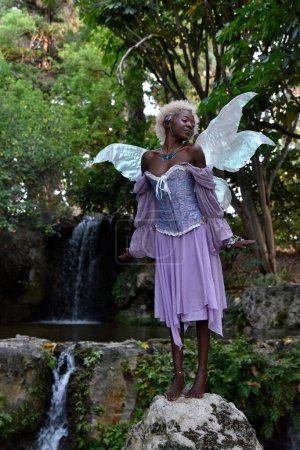 Téléchargez les photos : Portrait of beautiful African woman wearing purple fantasy costume, magical fairy wings and flower crown afro, wandering around forest location with natural lighting. - en image libre de droit
