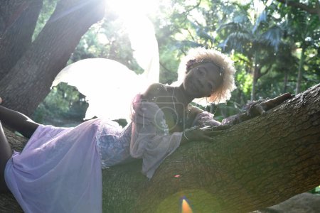 Téléchargez les photos : Portrait of beautiful African woman wearing purple fantasy costume, magical fairy wings & flower crown afro, climbing tree branches in forest location with natural glowing lighting. - en image libre de droit