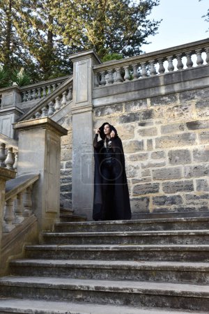 Foto de Portrait of beautiful female model with blonde plait, wearing black leather catsuit and flowing hooded cloak, fantasy assassin warrior.  Posing in castle background with stone staircase. - Imagen libre de derechos